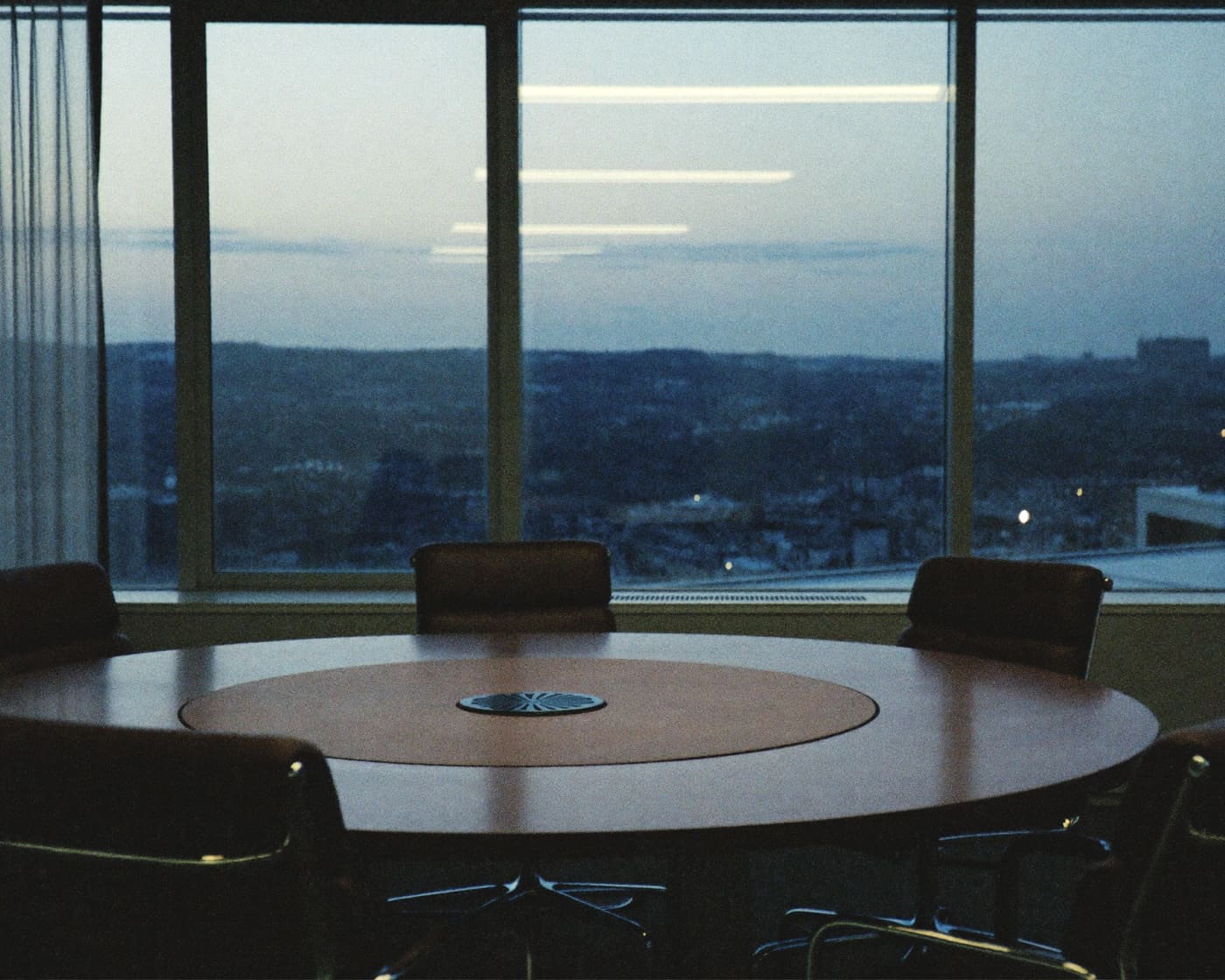 Round boardroom table by floor-to-ceiling windows overlooking a city at dusk