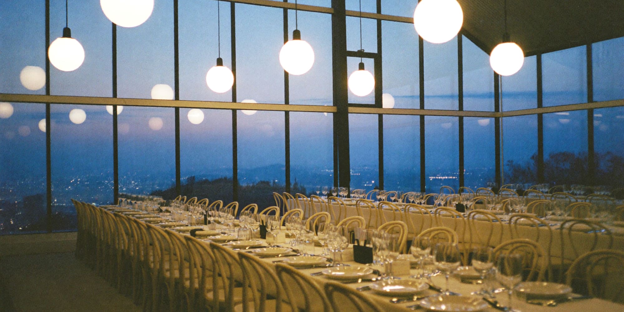 Corporate reception in a glass pavilion at dusk, banquet tables under warm pendant lights with a city view beyond